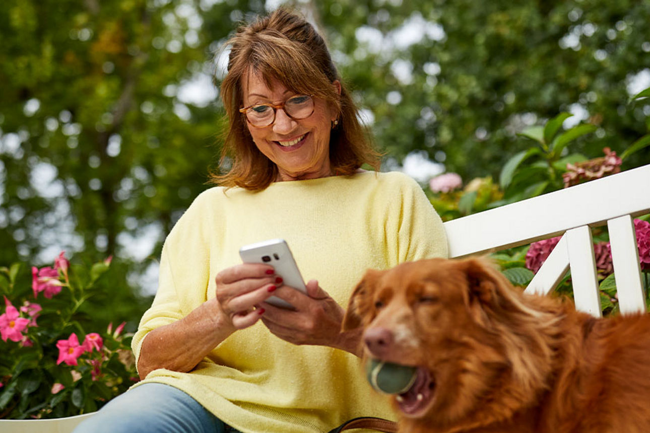 Frau ist am Handy und sitzt neben einem Hund auf einer Bank.