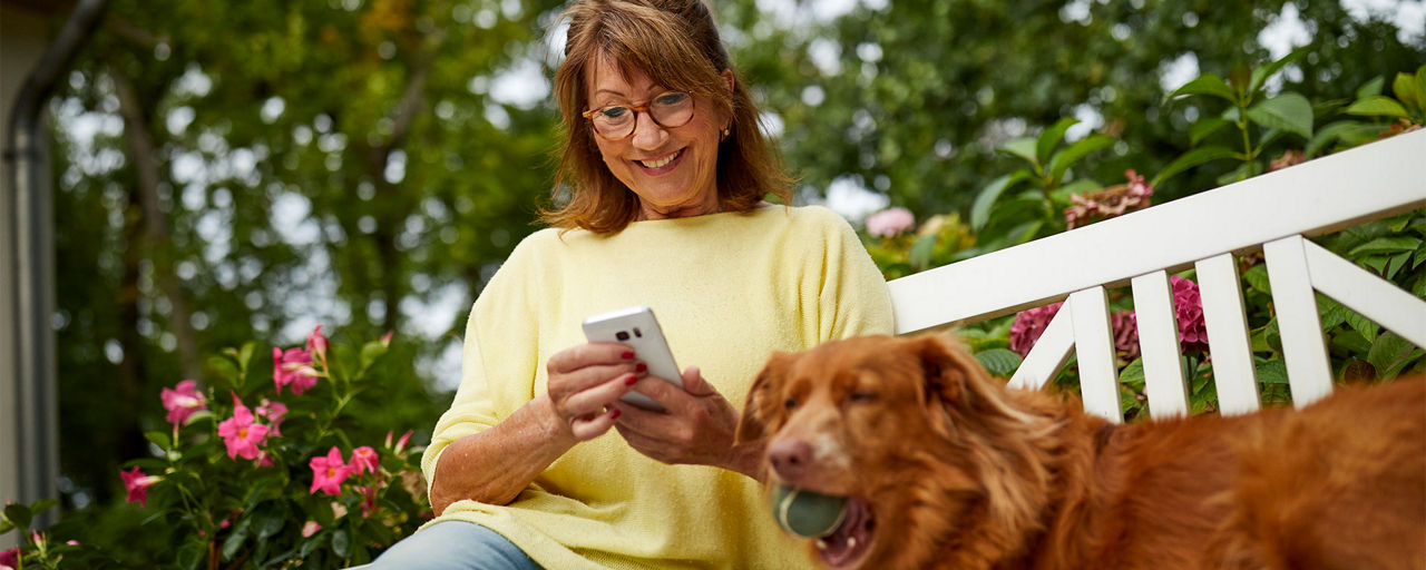 Frau mit Hund betreibt Banking am Smartphone auf einer Bank im Garten