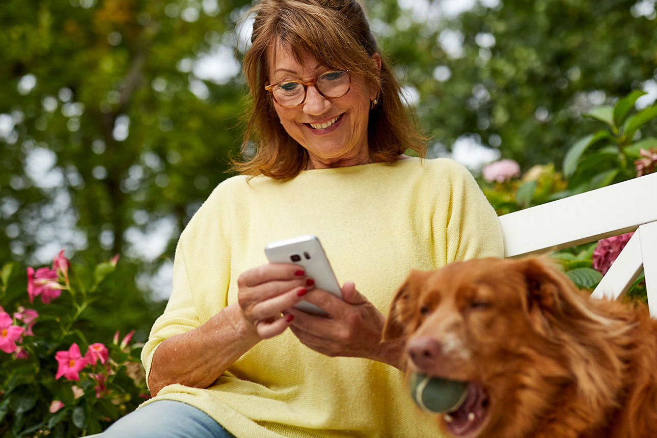 Frau mit Hund betreibt Banking am Smartphone auf einer Bank im Garten