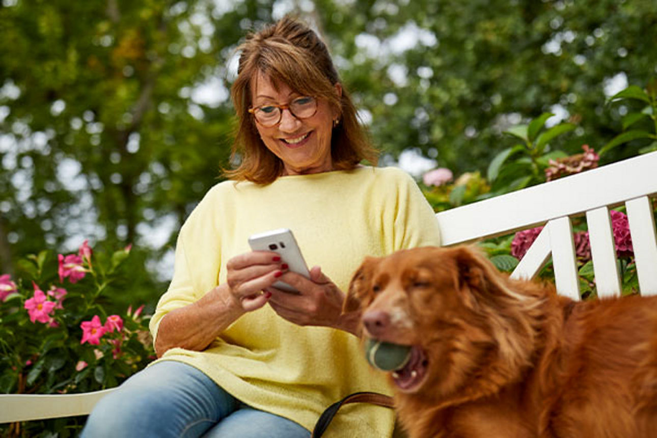 Frau schaut sitzend auf einer Bank auf ihr Handy mit ihrem Hund