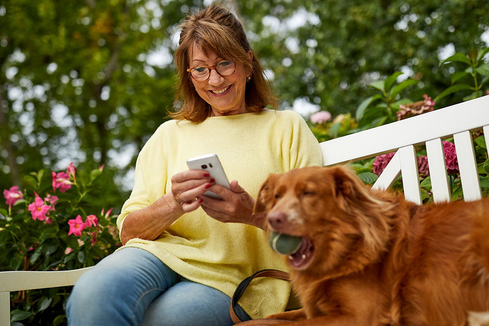 Frau mit Hund betreibt Banking am Smartphone auf einer Bank im Garten