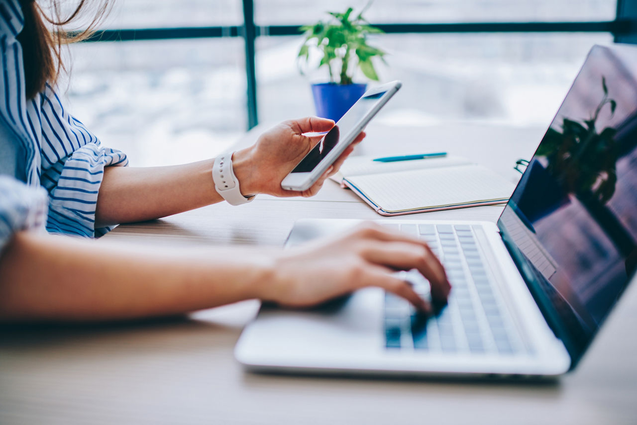 Hände einer Frau tippen auf einem Laptop, in einer Hand hält sie ein Smartphone