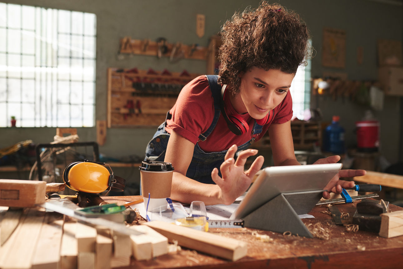 Eine Frau arbeitet in einer Holzwerkstatt an einer Werkbank und bedient konzentriert ein Tablet. Um sie herum liegen Holzstücke, Werkzeuge und Schutzutensilien.