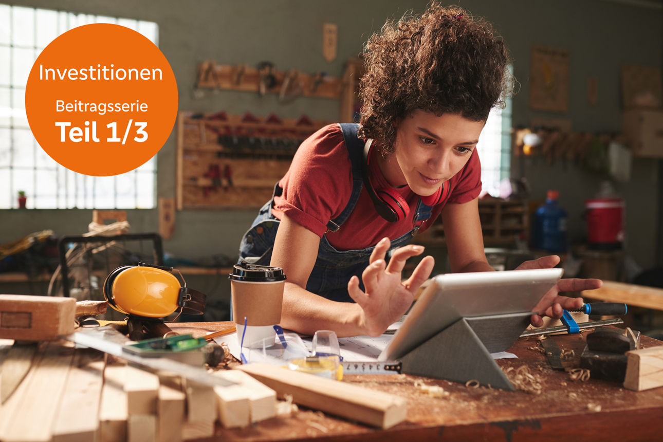 Investieren in die Zukunft Eine Frau arbeitet in einer Holzwerkstatt an einer Werkbank und bedient konzentriert ein Tablet. Um sie herum liegen Holzstücke, Werkzeuge und Schutzutensilien.