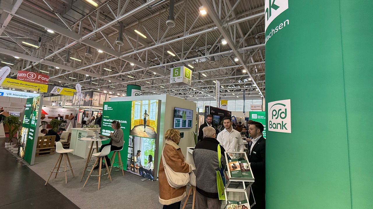 Seitlicher Blick auf den Messestand der PSD Bank München. Rechts ist eine große grüne Säule. Links daneben sind zwei Berater im Gespräch mit zwei Kunden. Mittig ist eine Leuchtrahmen zum Thema Baufinanzierung. Davor sitzt eine Beraterin und arbeitet.