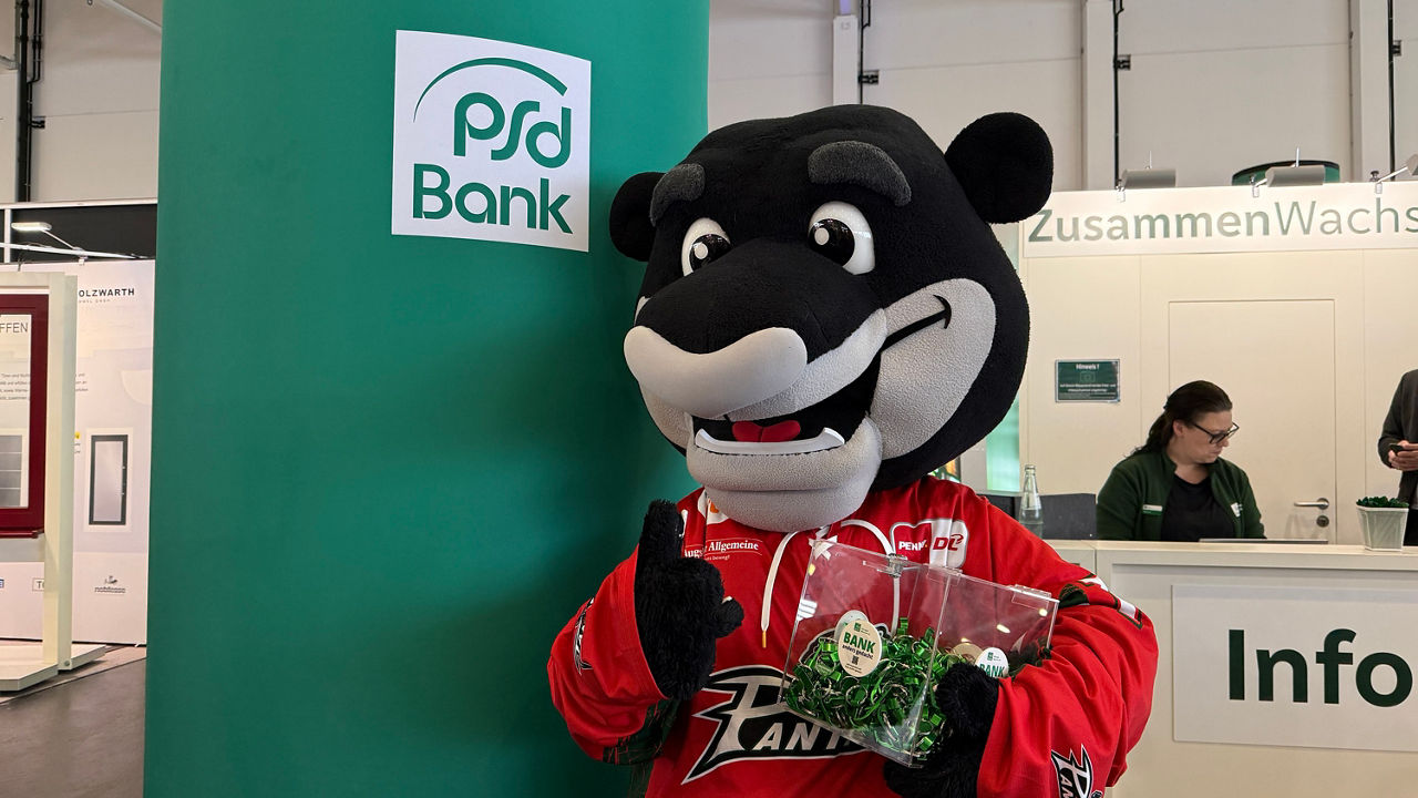 Im Vordergrund steht ein Panther im roten Trikot als Maskottchen. Es hält eine Box mit grünen Schlüsselanhängern in der liken Hand und zeigt mit der Rechten den Daumen hoch. Es steht vor einer grünen PSD Bank Leuchtsäule. Im Hintergrund ist die Info.