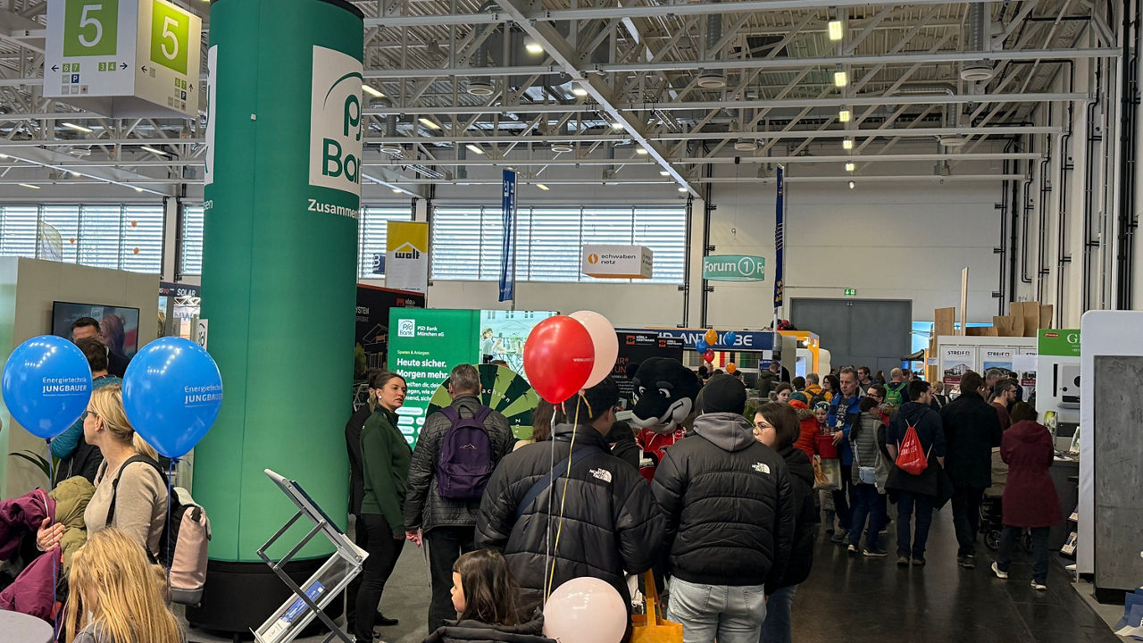 Seitlicher Blick auf den hinteren Bereich des Messestandes der PSD Bank München. Menschen tummeln sich vor dem Glücksrad. 