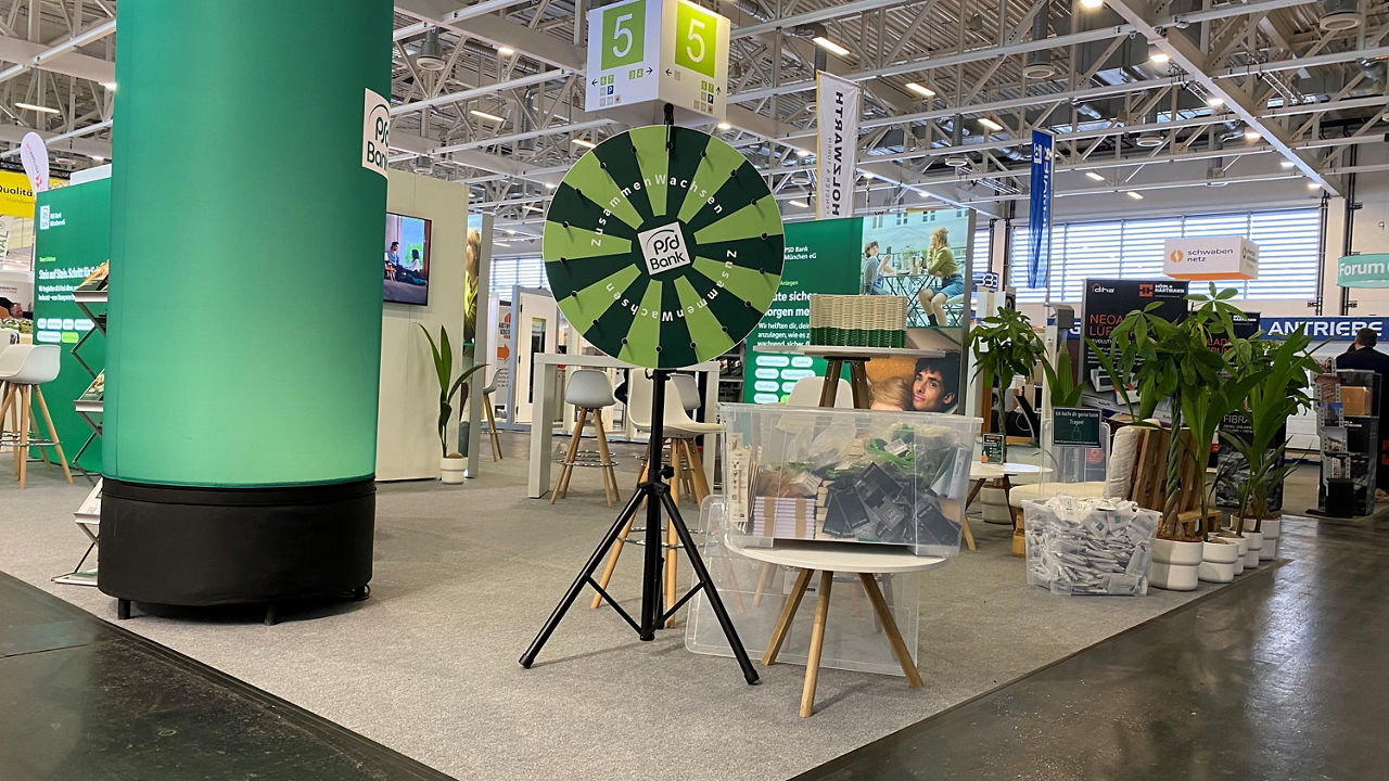Blick auf eine Ecke des Messestandes der PSD Bank München. Mittig im Vordergrund steht ein grün gestreiftes Glücksrad, daneben zwei Boxen mit Gewinnen. Links daneben ist eine grüne Leuchtsäule. Rechts im Bild ist eine Sitzecke zu erkennen. 