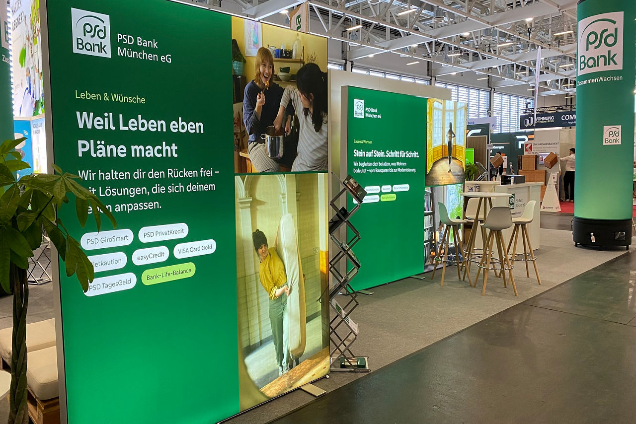 Seitlicher Blick auf den Messestand der PSD Bank München. Rechts ist eine große grüne Säule. Links daneben sind zwei Berater im Gespräch mit zwei Kunden. Mittig ist eine Leuchtrahmen zum Thema Baufinanzierung. Davor sitzt eine Beraterin und arbeitet.