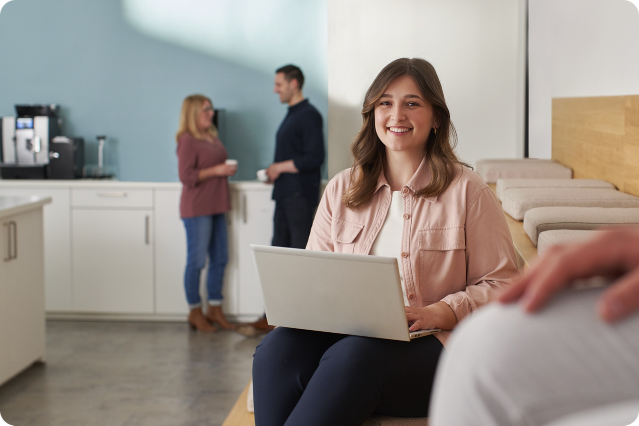 Eine junge Frau sitzt mit einem Laptop auf dem Schoß und lächelt in die Kamera, im Hintergrund unterhalten sich zwei Personen