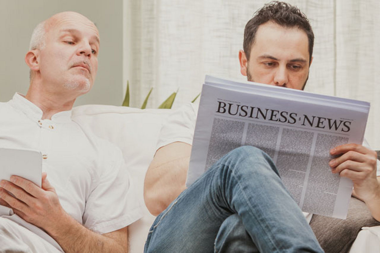 Zwei Männer sitzen auf dem Sofa, ein Mann liest Zeitung und der andere spickelt drauf