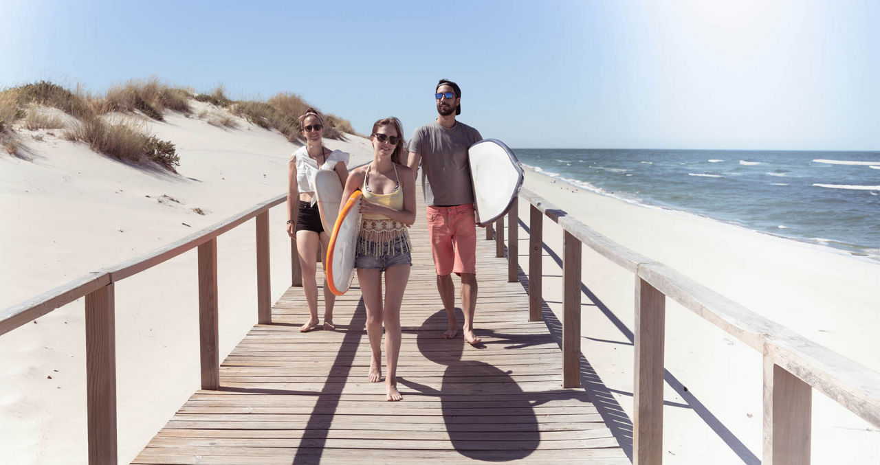 Boys and girls teen surfers with surfboards walking on a wooden pathway at the beach.