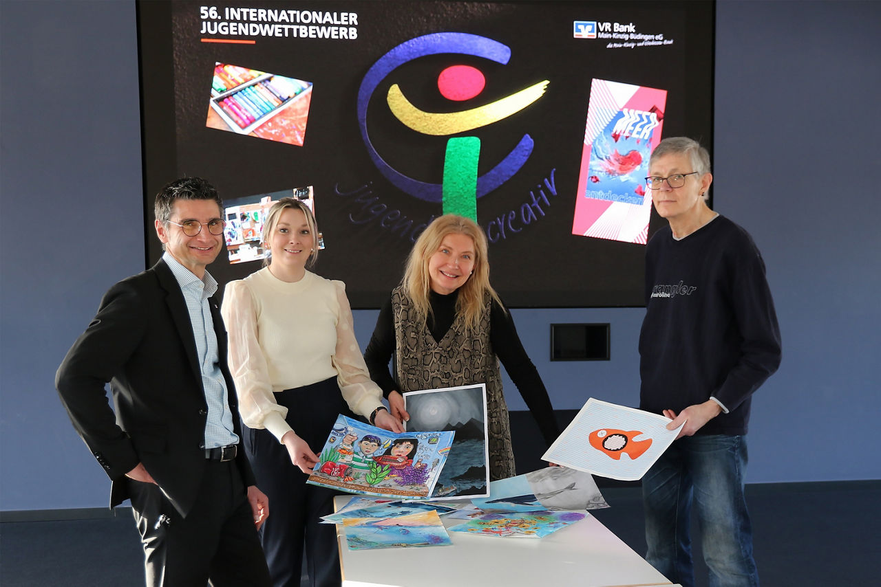 Vier Personen stehen hinter einem Tisch auf dem die Bilder der Teilnehmer liegen. Im Hintergrund sieht man das Logo von jugend creativ