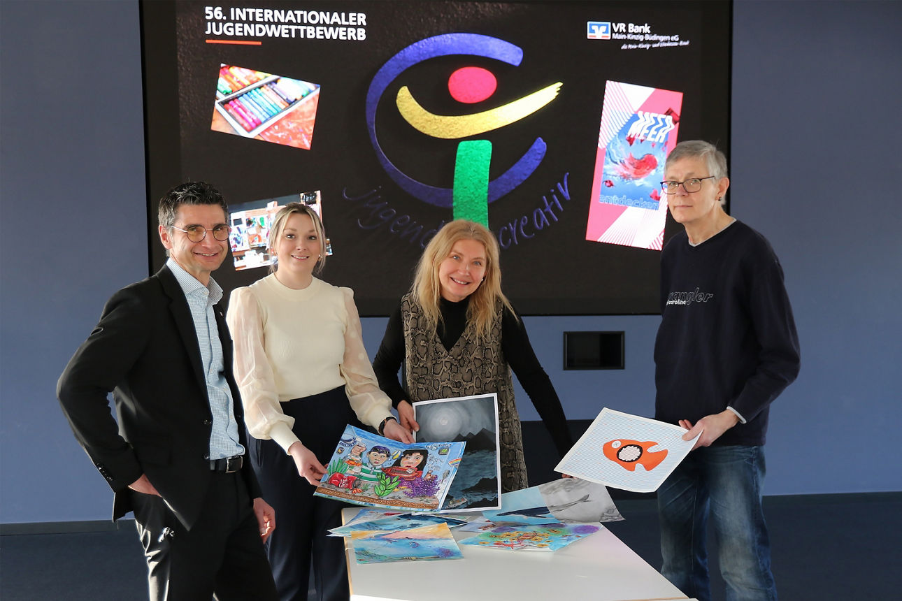 Vier Personen stehen hinter einem Tisch auf dem die Bilder der Teilnehmer liegen. Im Hintergrund sieht man das Logo von jugend creativ