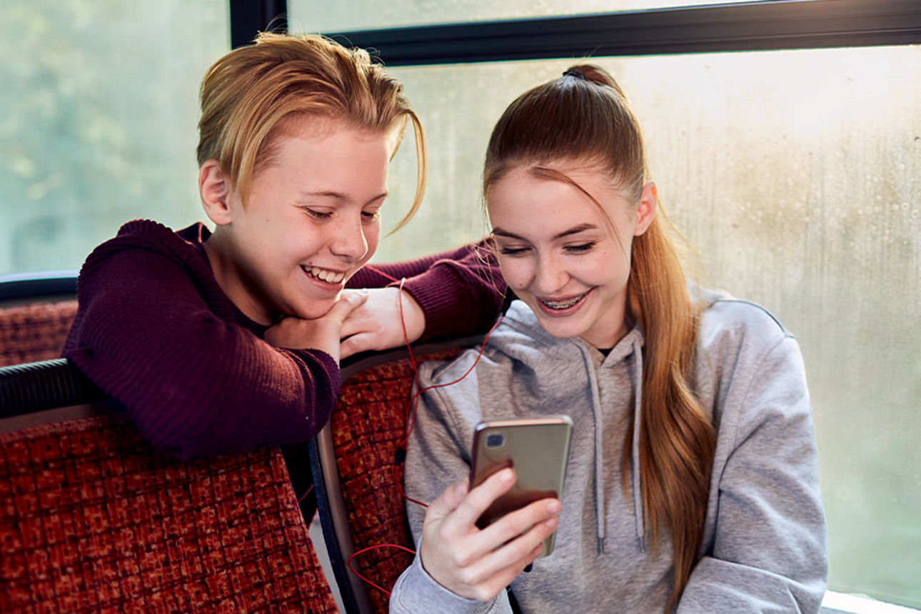 Ein Mädchen und ein Junge sitzen im Bus und schauen auf ein Smartphone.