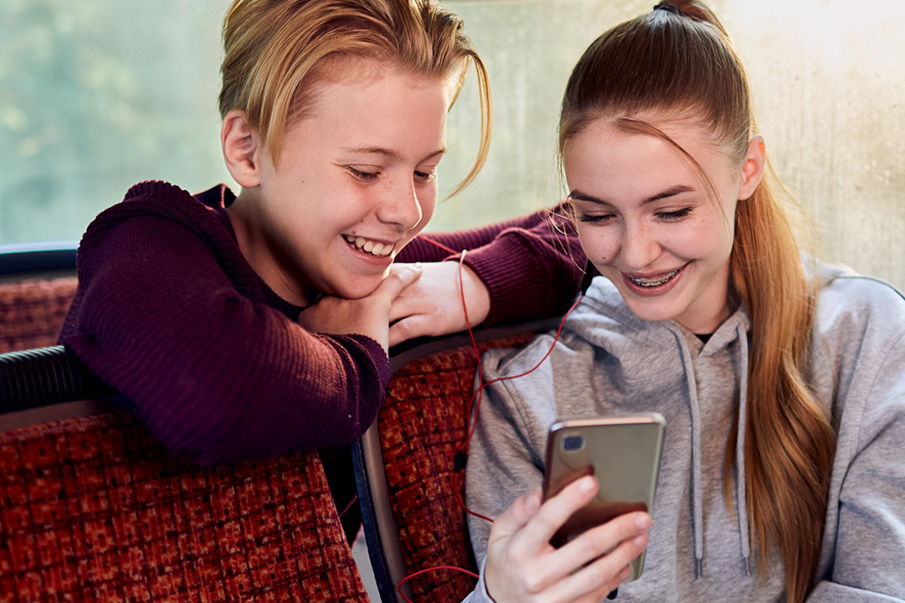 Zwei Jugendliche sitzen im Bus und schauen lächelnd in ein Smartphone