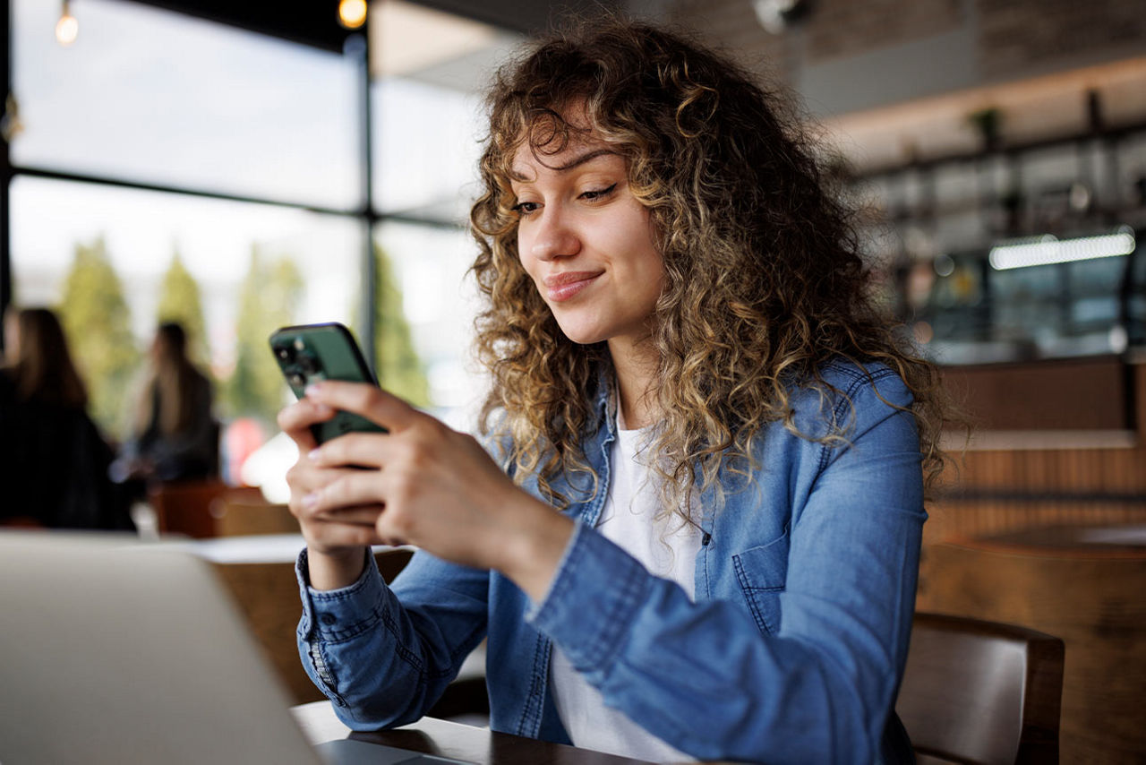 Junge Frau sitzt in einem Café vor ihrem Laptop und tippt auf ihrem Smartphone