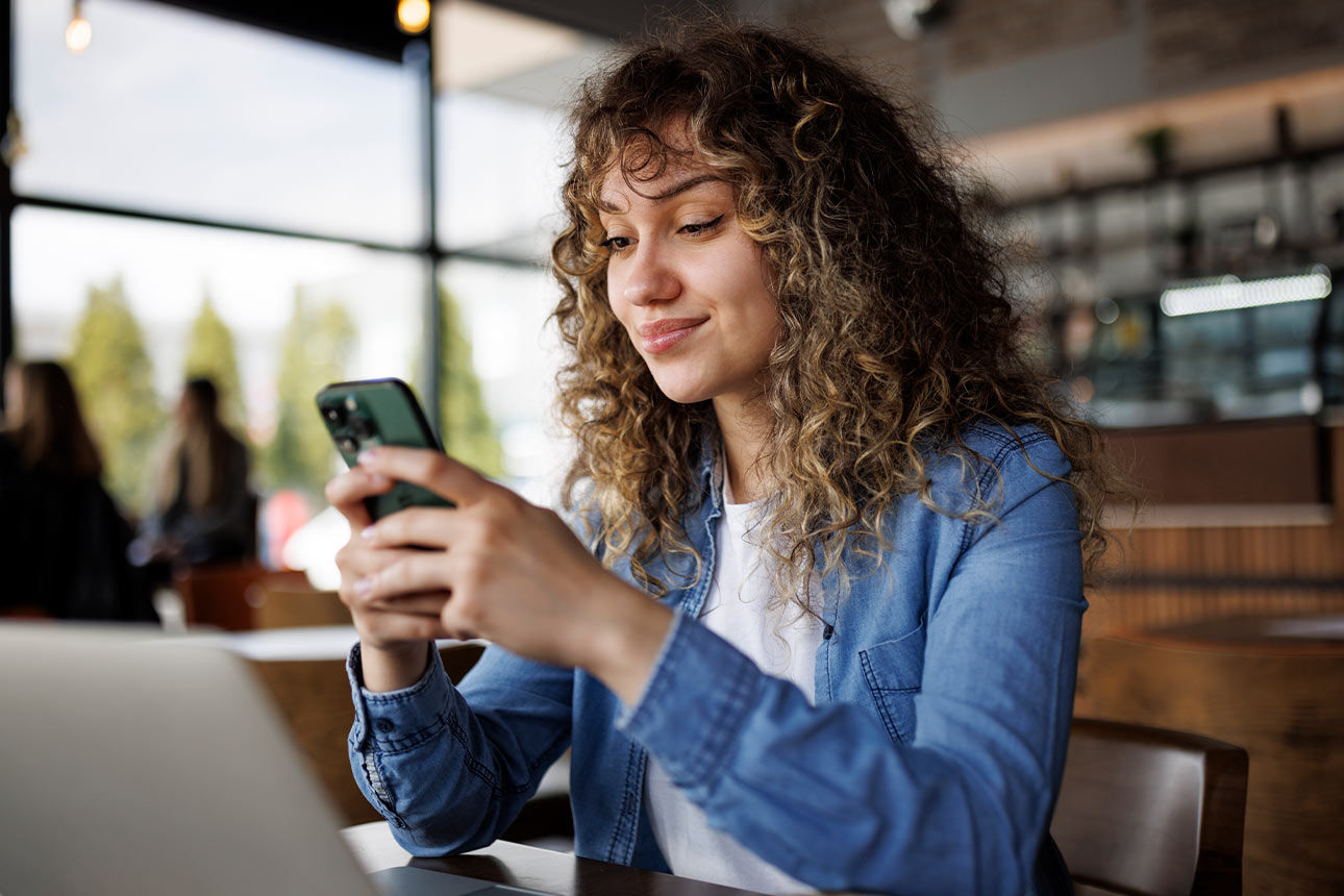 Junge Frau sitzt in einem Café vor ihrem Laptop und tippt auf ihrem Smartphone