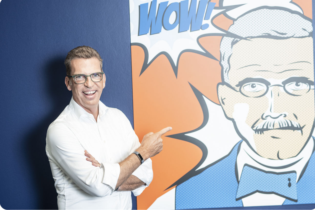 Carsten Jung steht vor einem Schild mit einer Illustration von Raiffeisen und dem Wort Wow und zeigt lachend darauf