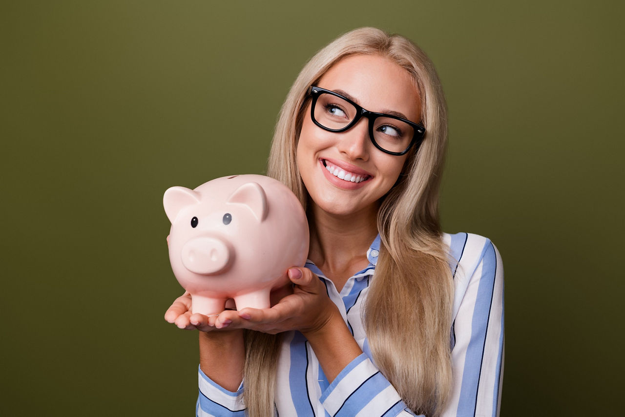 Eine blonde Frau mit Schwarzer Brille trägt ein Sparschwein in ihren Händen und lächelt dabei.