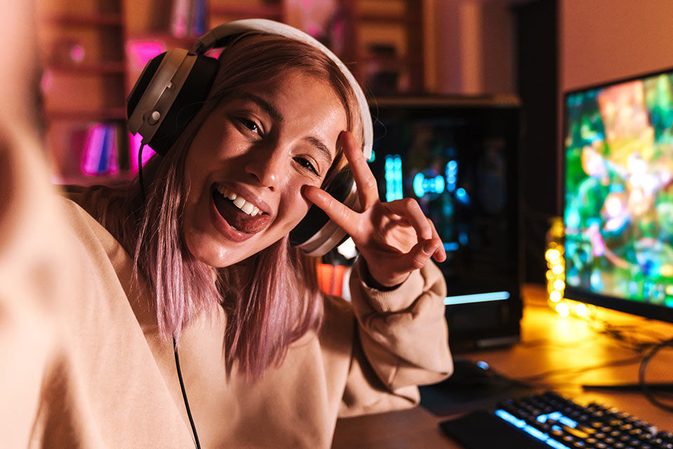 Ein junges Mädchen sitzt grinsend mit Headset vor einem PC und macht ein Victory-Zeichen in die Kamera.
