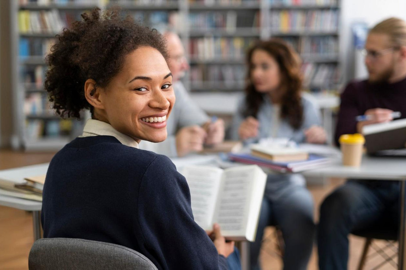 Bild von mehreren jungen Leuten, die in einer Bibliothek sitzen. Eine junge Frau mit einem Buch blickt in die Kamera.