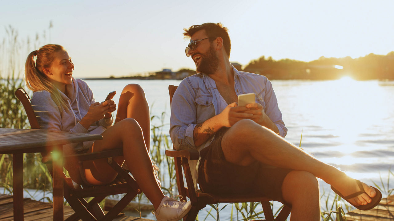 Bild von zwei Personen, die an einem See sitzen und gemeinsam lachen. Beide halten jeweils ein Handy in den Händen.