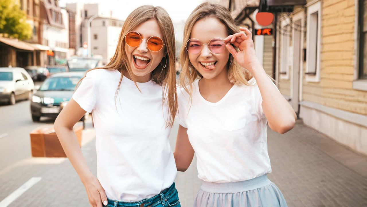 Bild von zwei jungen Frauen mit orangenen Sonnenbrille, die lächelnd und frech in die Kamera blicken.