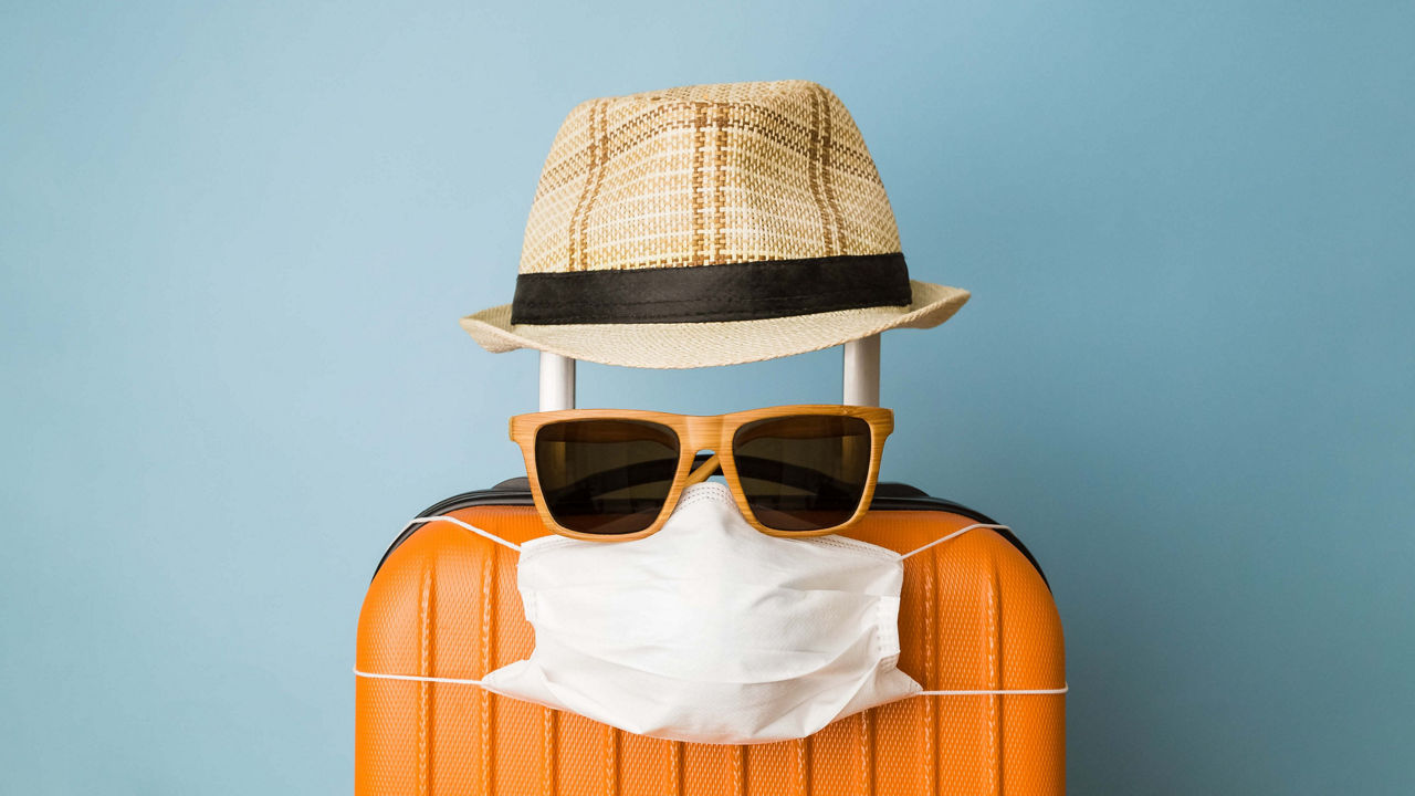 Bild von einem orangenen Koffer, der mit einem Hut, einer Sonnenbrille und einer Gesichtsmaske dekoriert ist.