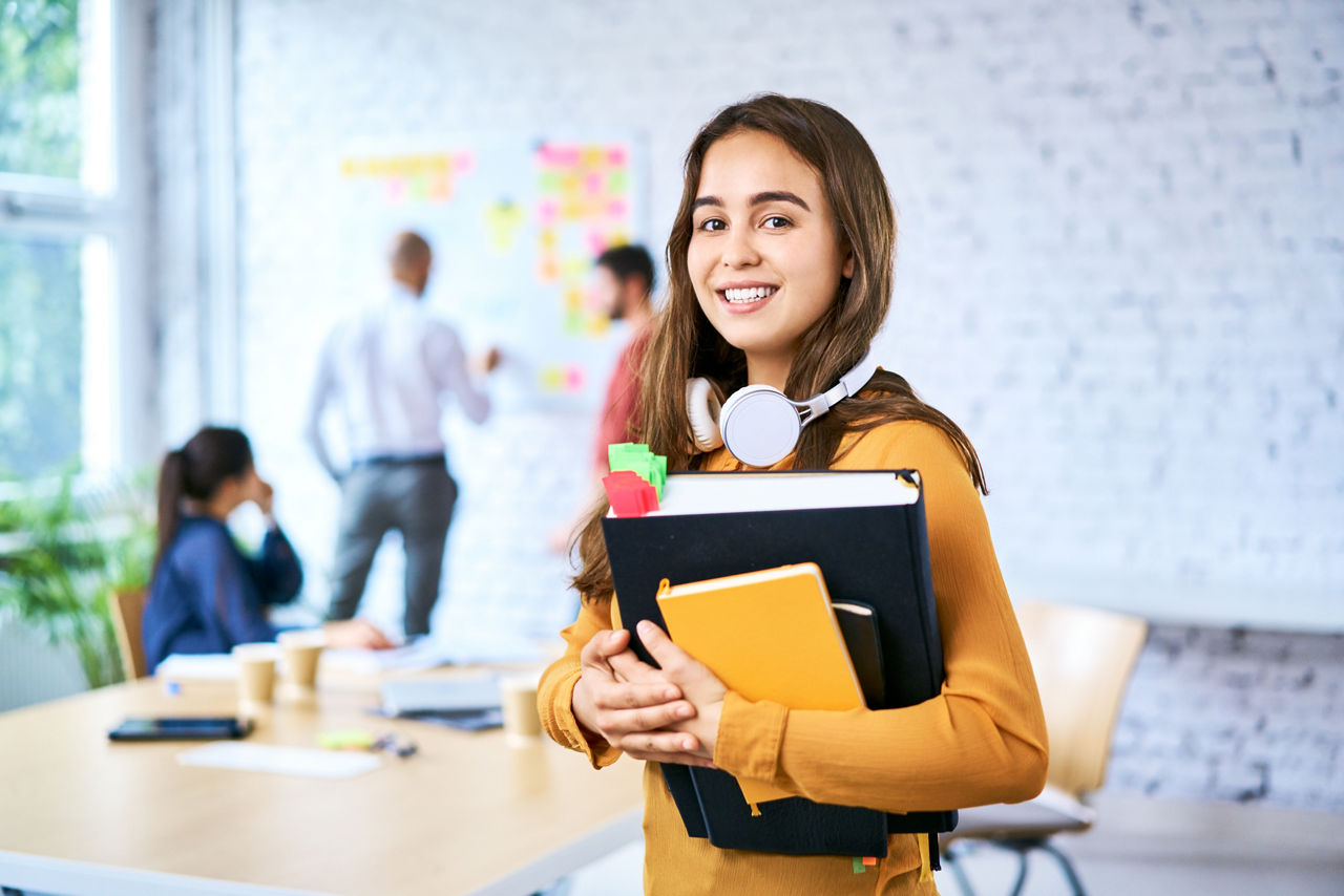 Junge Frau mit Kopfhörern, Ordnern und Notizbuch lächelt in einem modernen Büro