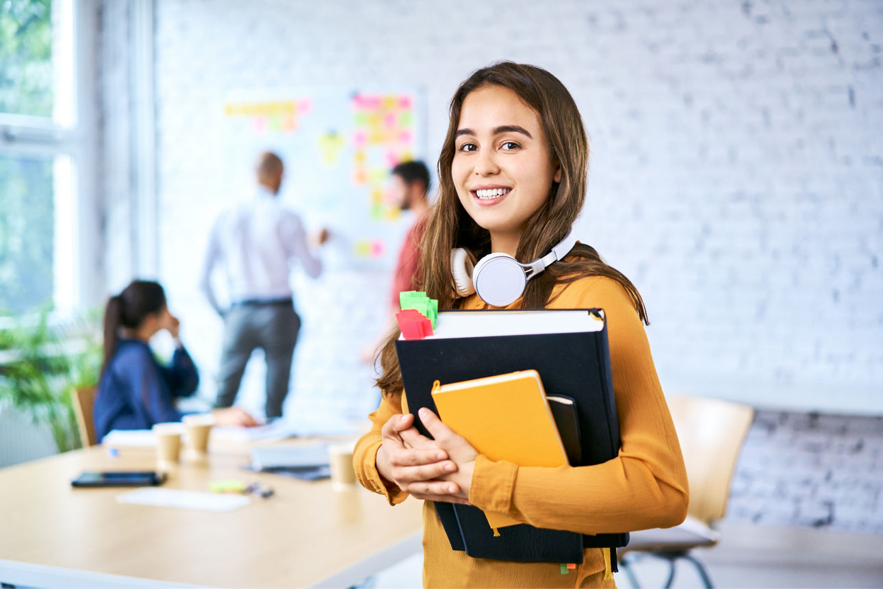 Junge Frau mit Kopfhörern, Ordnern und Notizbuch lächelt in einem modernen Büro