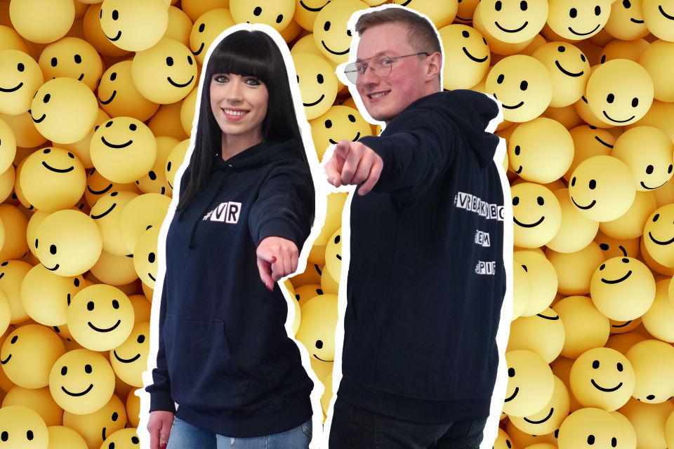 Zwei junge Menschen vor einer Wand aus Smileys deuten auf den Betrachter