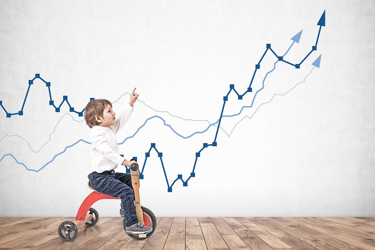 Kleines Kind auf einem Dreirad vor einer Wand mit einem abgebildeten Aktien-Chart.