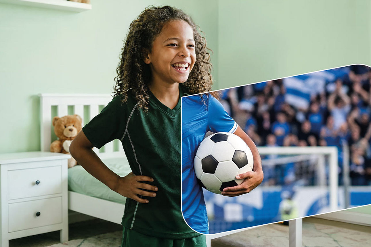 Ein lachendes dunkelhäutiges Mädchen im Trikot lacht mit Fußball in der Hand im Kinderzimmer.
