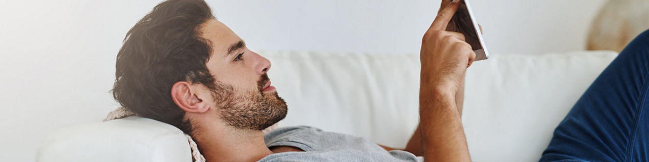 Mann mit dunkelbraunen Haaren liegt auf dem Sofa mit dem Handy in der Hand