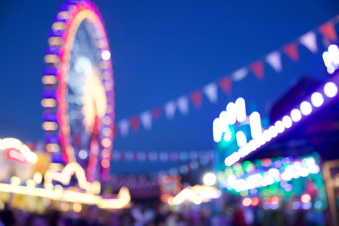 Unscharfe Aufnahme eines Riesenrads und beleuchteter Fahrgeschäfte auf einem Jahrmarkt bei Abenddämmerung.