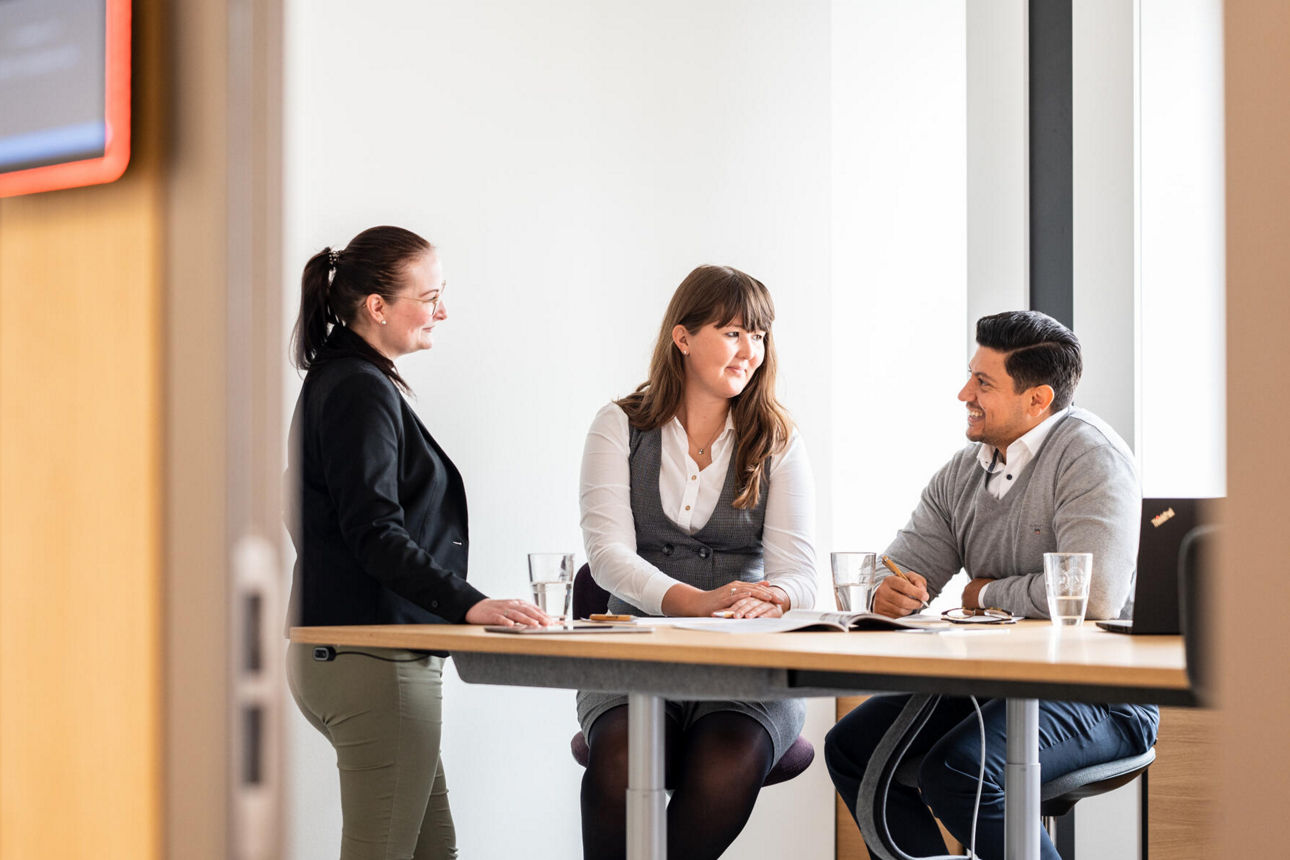 Zwei Mitarbeiterinnen und ein Mitarbeiter besprechen sich stehend am einem Tisch