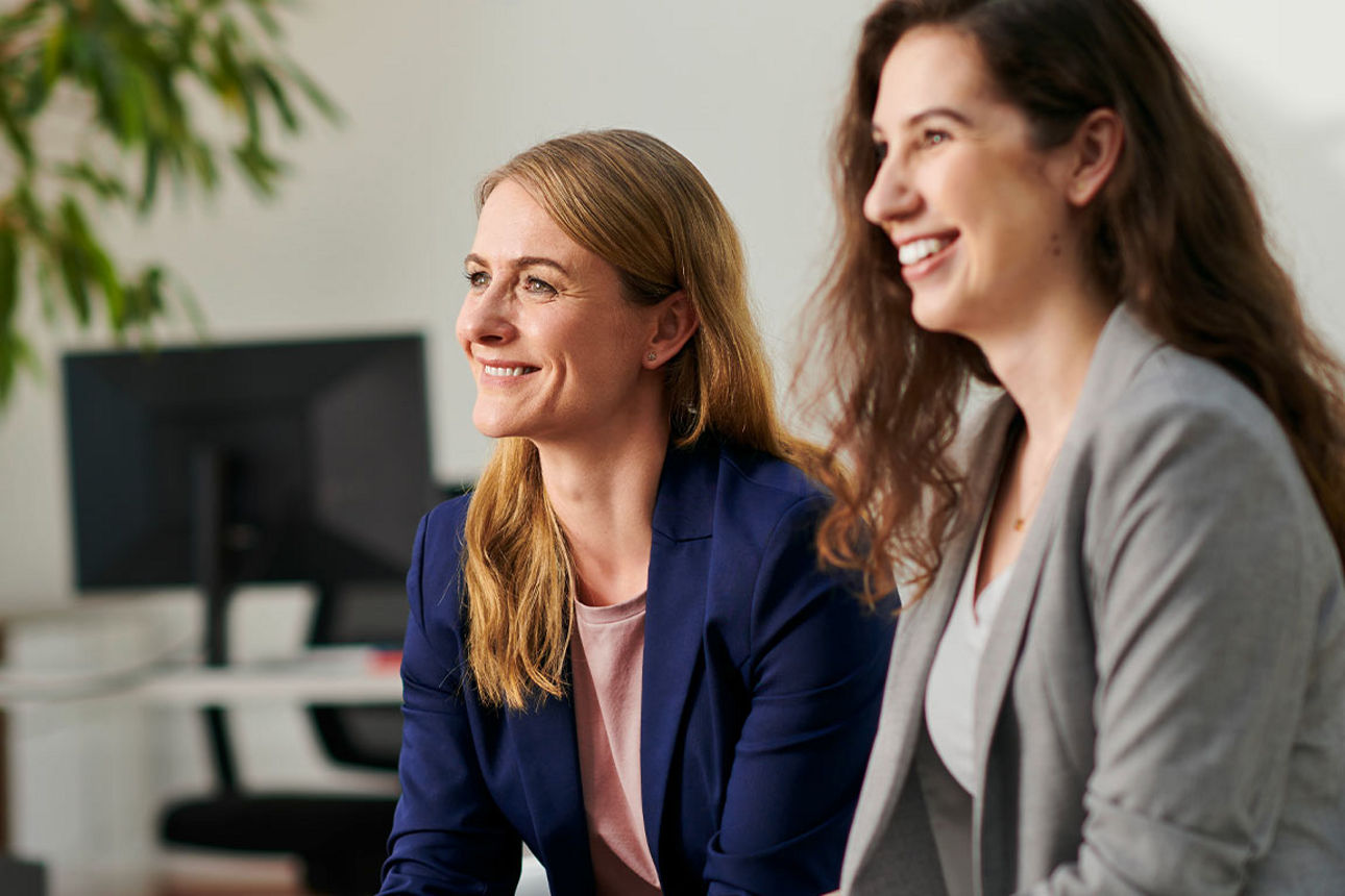 Zwei lächelnde Frauen sitzen nebeneinander in einem Büro