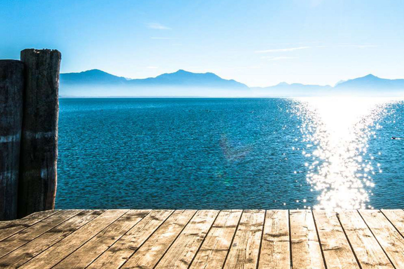 Holzsteg am See mit glitzerndem Sonnenlicht und Bergpanorama im Hintergrund