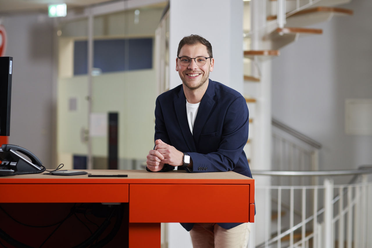 Ein junger Mann mit Brille stützt sich stehend auf einen Schreibtisch und lächelt in die Kamera