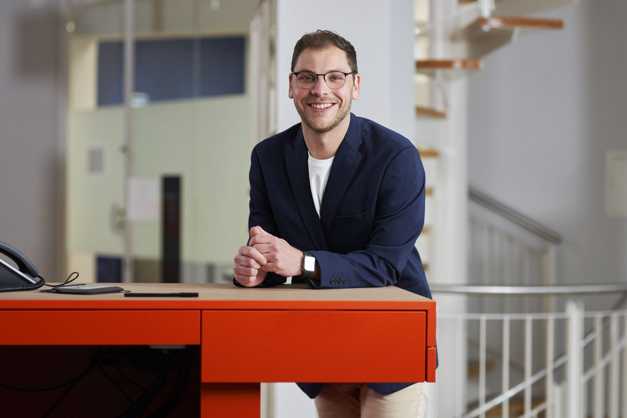 Ein junger Mann mit Brille stützt sich stehend auf einen Schreibtisch und lächelt in die Kamera