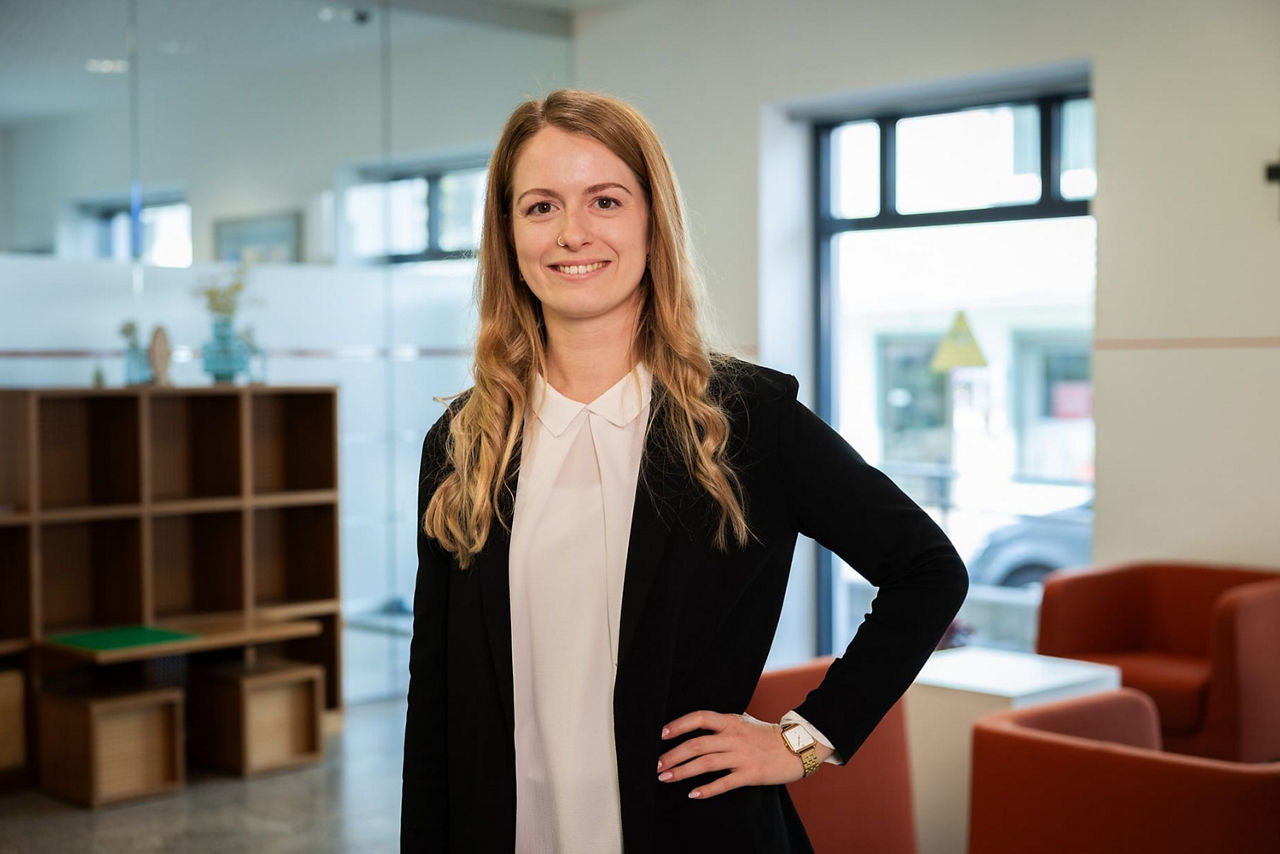 Professionelle Frau in Blazer und weißer Bluse mit Hand in der Hüfte. Freundliches Lächeln im modernen Büro.