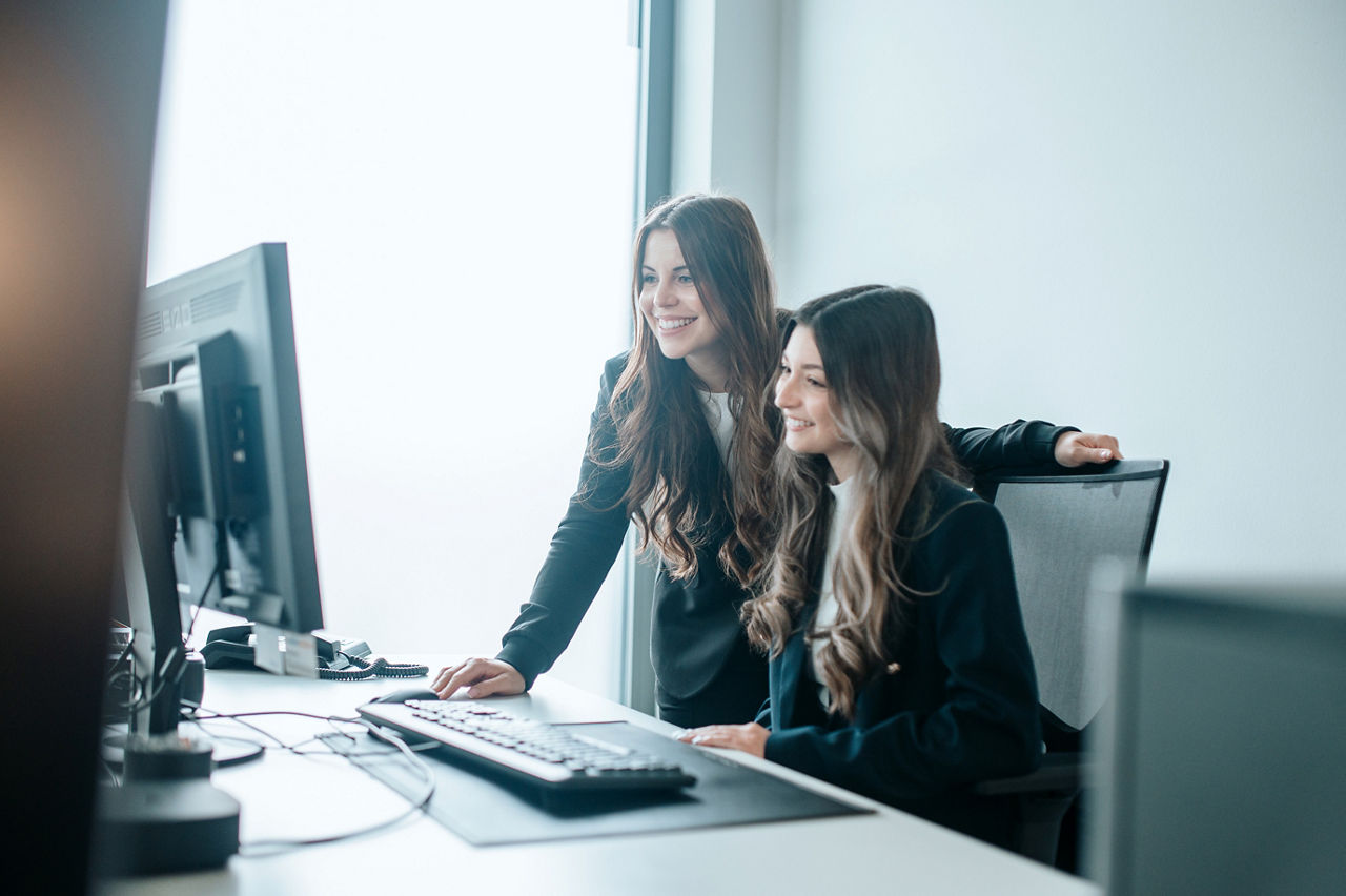 Zwei Frauen sitzen am Schreibtisch und schauen lächelnd auf einen Computer.