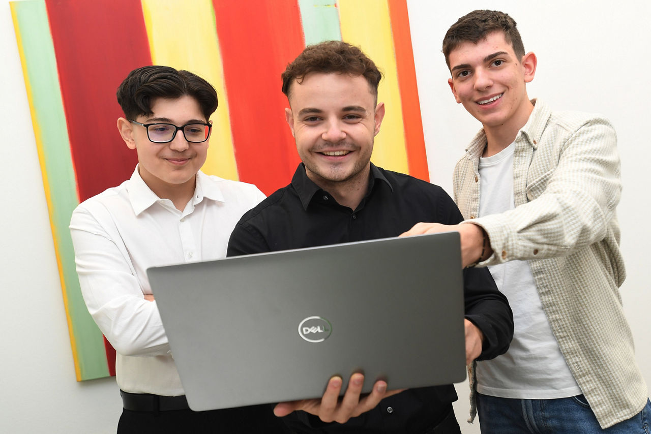 Drei relativ junge Männer stehen nebeneinander in einem Büro. Der Mann in der Mitte hält einen geöffneten Laptop in den Händen. Der Mann links von ihm zeigt etwas am Bildschirm. Der Mann rechts von ihm blickt in den Laptop.