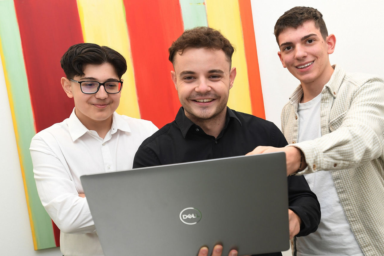 Drei relativ junge Männer stehen nebeneinander in einem Büro. Der Mann in der Mitte hält einen geöffneten Laptop in den Händen. Der Mann links von ihm zeigt etwas am Bildschirm. Der Mann rechts von ihm blickt in den Laptop.