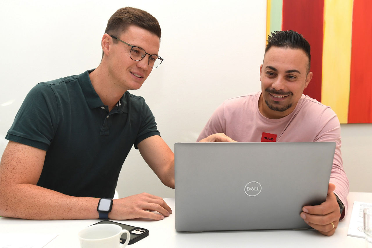 Zwei Männer sitzen in einem Büro am Schreibtisch und blicken in den Laptop. Der Mann mit Brille erklärt seinem Kollegen links von ihm etwas.