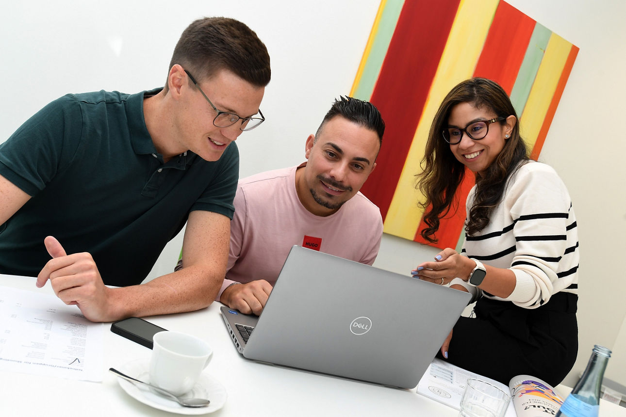 Zwei Männer sitzen in einem Büro am Schreibtisch und blicken in den Laptop. Eine Frau sitzt zur halb auf dem Schreibtisch und deutet erklärend in den Bildschirm des Laptops.
