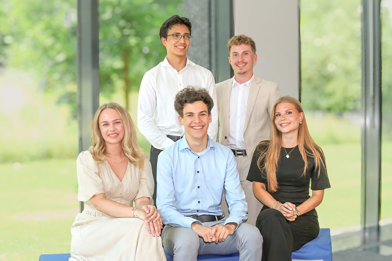 Fünf Auszubildende sitzen und stehen für ein Gruppenfoto vor großen Fenstern.