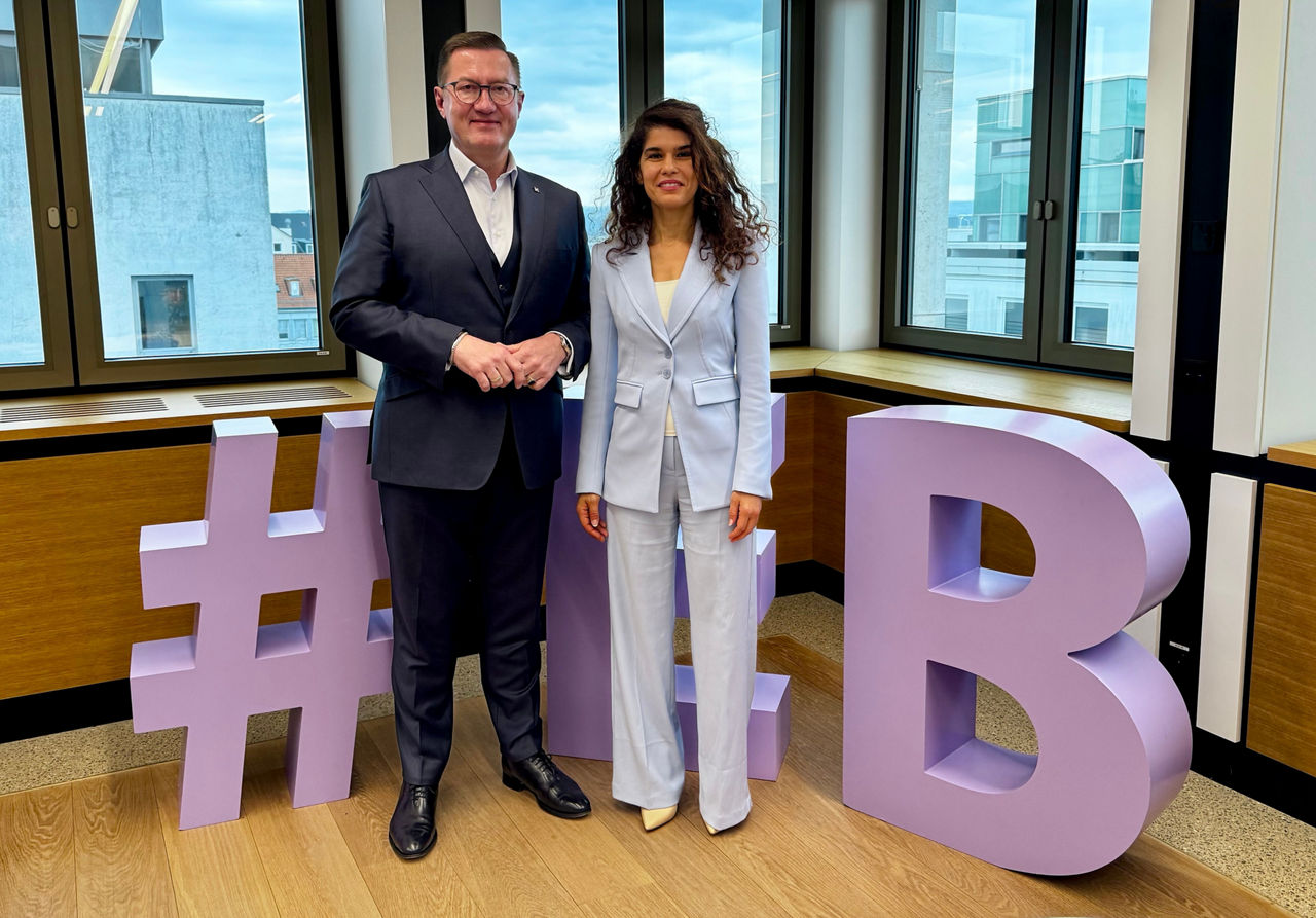 Thomas Katzenmayer und Souad Benkredda stehen in einem hellen Besprechungsraum vor großen Fenstern in der Evangelischen Bank.