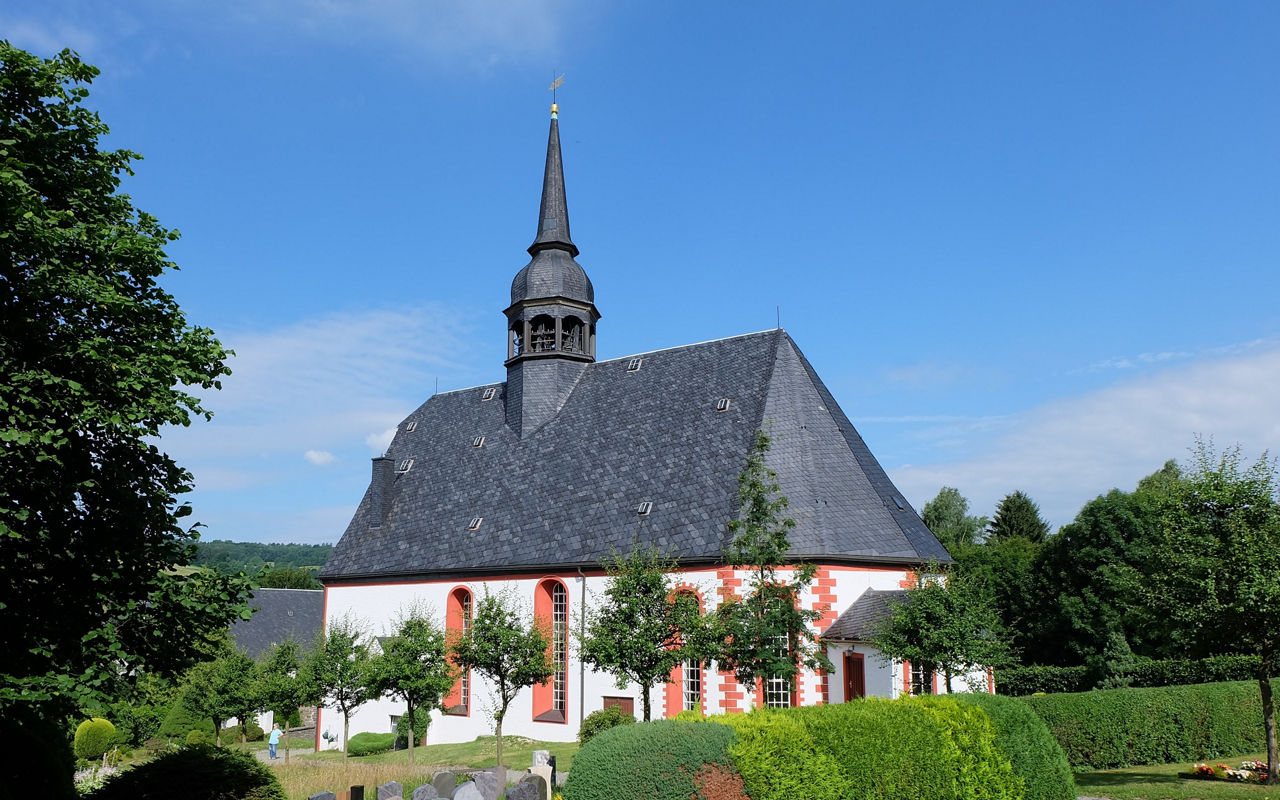Eine Kirche steht auf einem Hügel und im Hintergrund sieht man grüne Wiesen und Windräder
